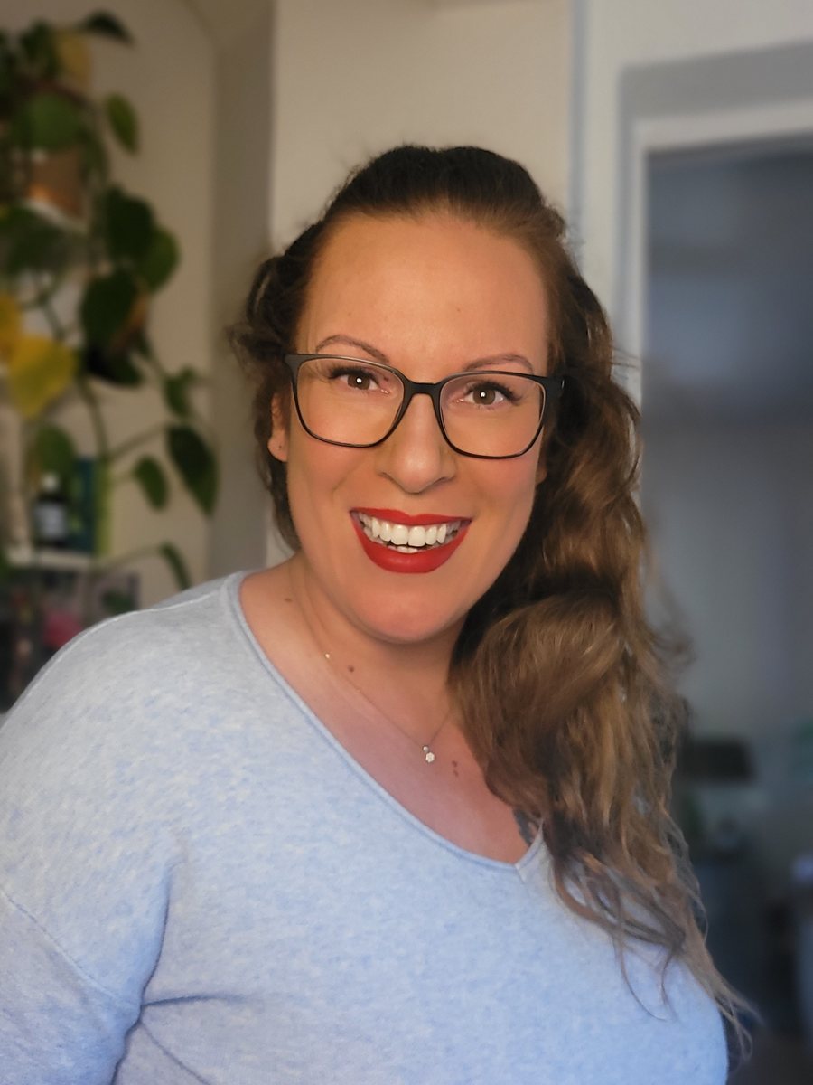 Alexandra Georg lacht in die Kamera, mit Brille, rotem Lippenstift und hellblauem Pullover